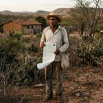 otografia de um produtor rural idoso, de pele morena e chapéu de palha, em pé no centro de uma paisagem seca da Caatinga no Ceará. Ele veste camisa clara de mangas longas e calça comprida, expressando preocupação ao segurar um documento oficial impresso (auto de infração). Ao fundo, vê-se uma casa de taipa simples, vegetação nativa com cactos (mandacarus), arbustos secos e, ao longe, uma caminhonete oficial de fiscalização ambiental estacionada perto das colinas sob o céu de fim de tarde