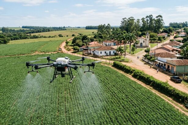 Fotografia aérea em ângulo plongée mostrando um grande drone agrícola cinza sobrevoando e pulverizando uma plantação verde e densa. O drone está em primeiro plano, à esquerda, liberando uma névoa fina sobre as culturas. Ao fundo, à direita, há uma pequena e charmosa vila rural com uma estrada de terra, casas de alvenaria com telhados de barro, uma pequena igreja branca com torre sineira e palmeiras, cercada por colinas e vegetação sob um céu azul com nuvens esparsas